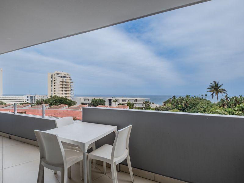 A spacious covered balcony featuring tiled flooring and a modern glass balustrade, offering expansive views of the surrounding neighborhood and the ocean.