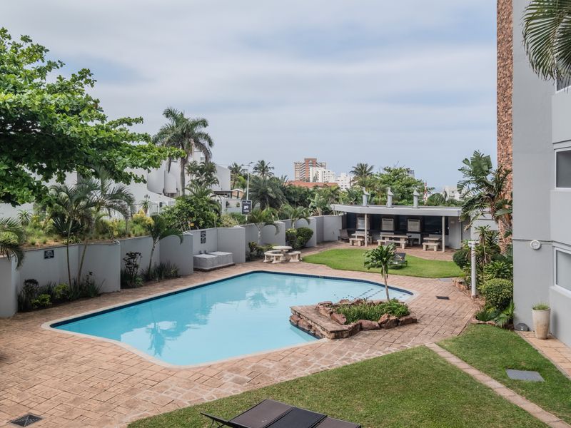 A well-maintained communal pool area featuring a paved deck, landscaped garden, and a covered outdoor braai facility for residents.
