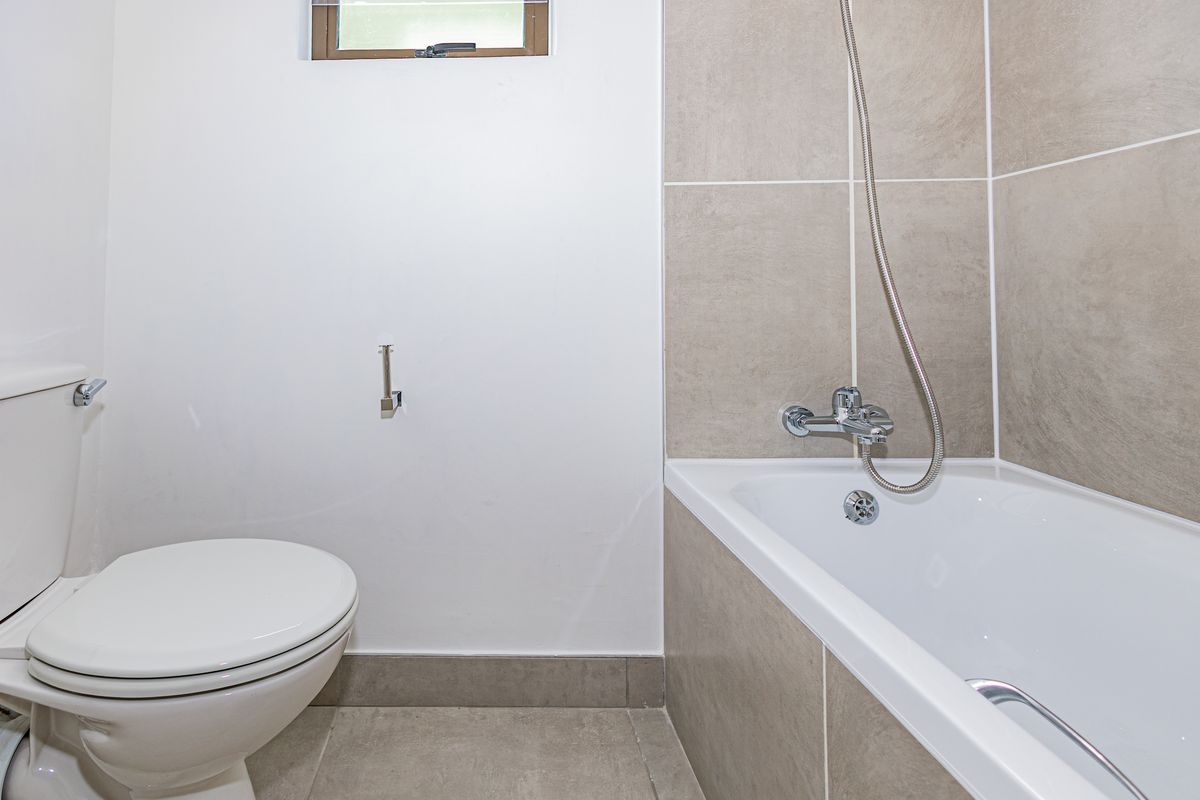 A clean, functional bathroom featuring a white toilet, a bathtub with a shower mixer, and neutral-toned wall and floor tiling.