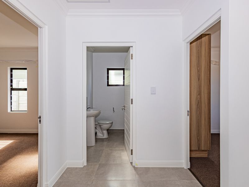 A bright, neutral hallway featuring tiled flooring, leading to a bathroom with a pedestal basin and toilet, and adjacent bedroom access.