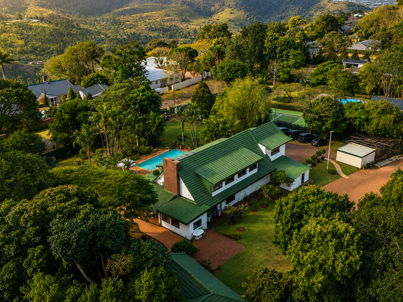 Aerial view of a residential property featuring a green tiled roof, swimming pool, landscaped garden, and paved driveway set within a lush green environment.