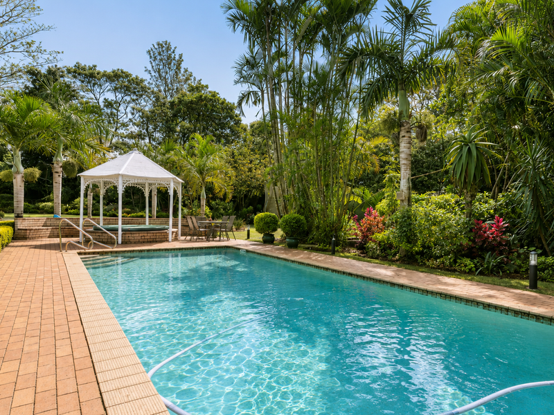 A well-maintained outdoor pool area featuring brick paving, a decorative gazebo, and lush tropical landscaping.
