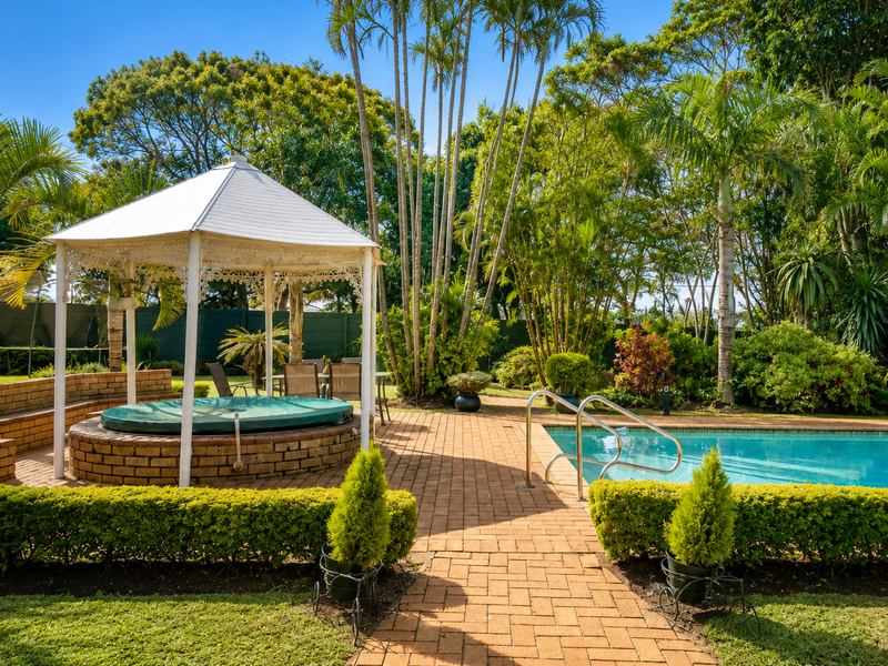 A well-maintained outdoor pool area featuring a swimming pool, a raised spa with a gazebo, and brick-paved surrounds set within a lush garden.