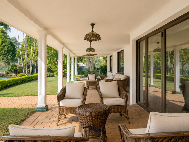 A spacious covered patio featuring paved flooring, decorative columns, and large sliding glass doors, overlooking a well-maintained garden and pool area.