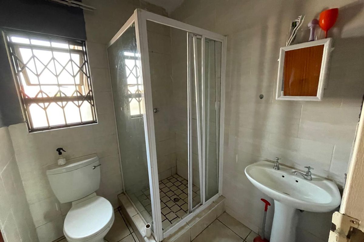 A functional bathroom featuring a shower enclosure, pedestal basin, and toilet, with tiled finishes and security bars on the window.