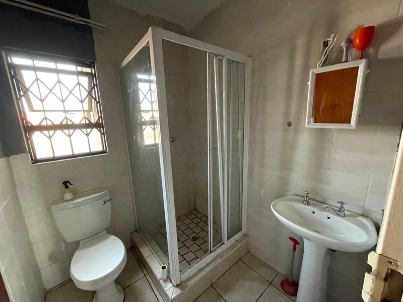 A functional bathroom featuring a shower enclosure, pedestal basin, and toilet, with tiled finishes and security bars on the window.