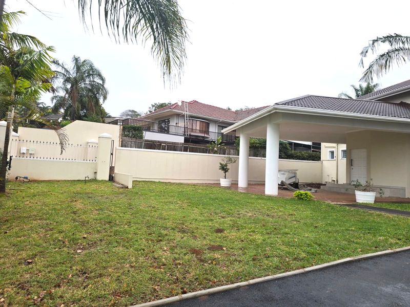 Front view of the property featuring a covered patio area, paved driveway, and a perimeter wall with a well-maintained garden space.