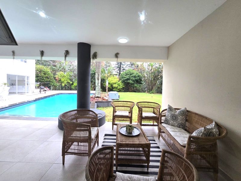 A spacious covered patio area featuring tiled flooring and recessed lighting, overlooking a private swimming pool and landscaped garden.
