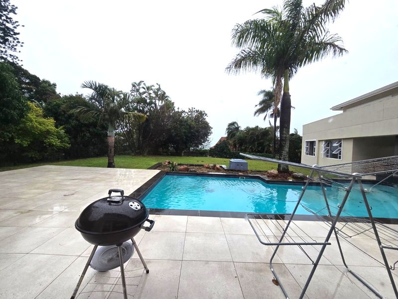 A well-maintained outdoor pool area featuring a tiled patio, swimming pool, and surrounding landscaped garden with mature palm trees.