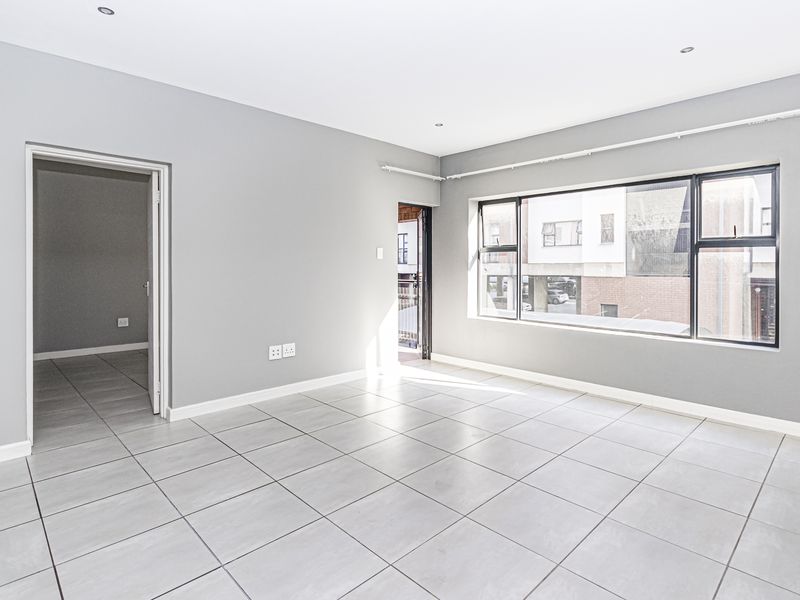 A bright, empty living area featuring neutral grey walls, large aluminium-framed windows, and modern tiled flooring, offering a clean and versatile space.