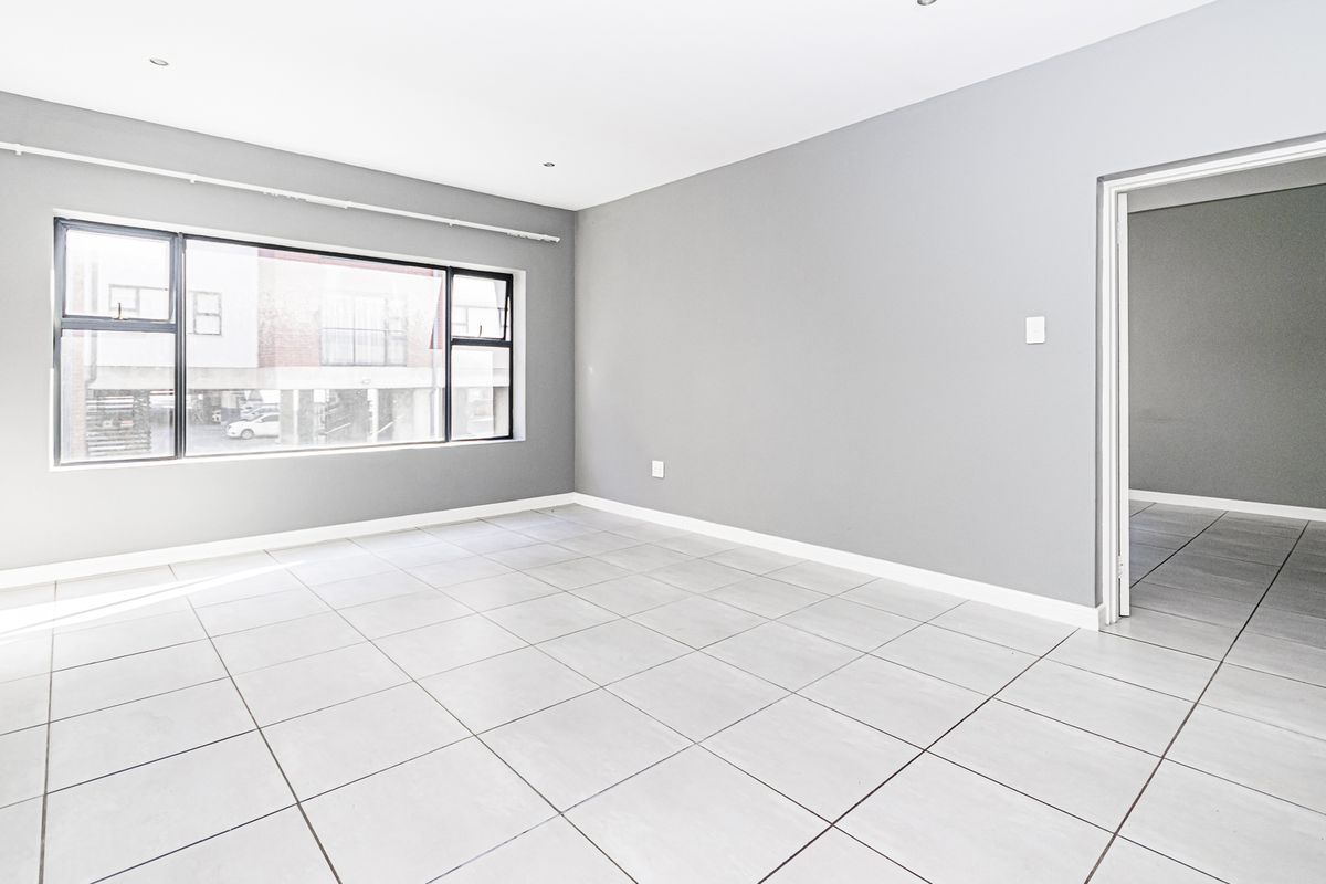 A bright, empty living area featuring neutral grey walls, large aluminium-framed windows, and clean tiled flooring throughout.