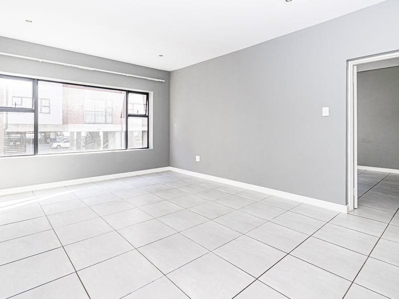 A bright, empty living area featuring neutral grey walls, large aluminium-framed windows, and clean tiled flooring throughout.