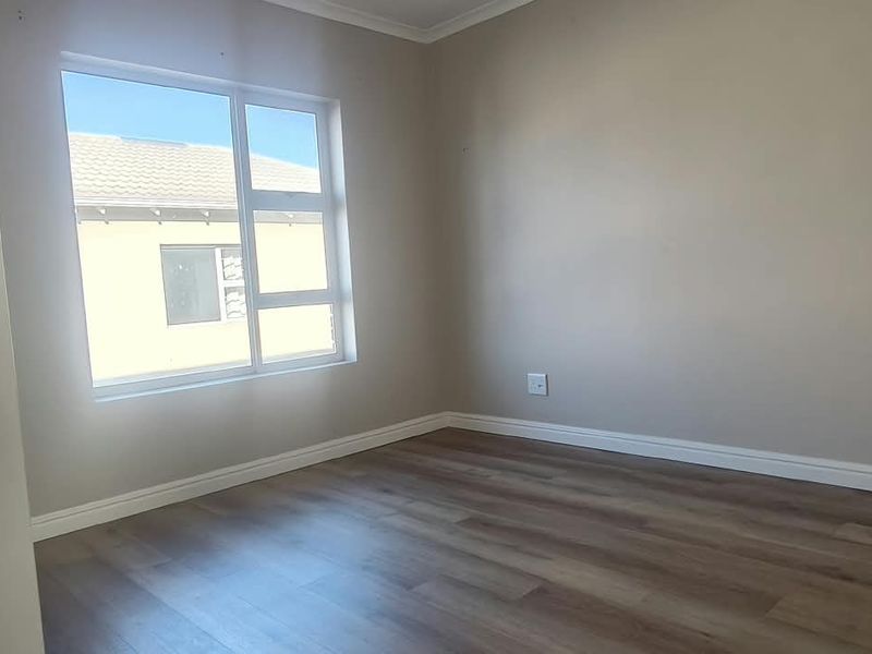 A bright, empty bedroom featuring modern laminate flooring, neutral wall tones, and a large aluminium-framed window providing natural light.