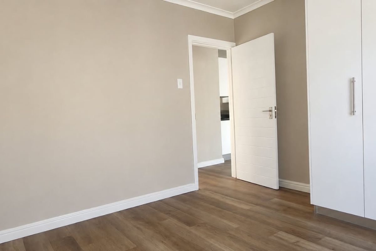 A bright, empty bedroom featuring modern laminate flooring, neutral wall tones, and white built-in cupboards with sleek hardware.
