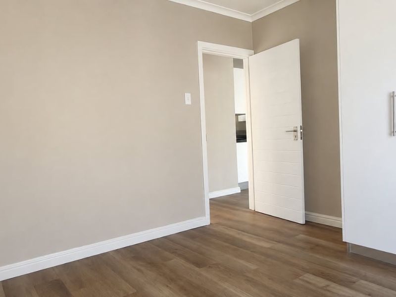 A bright, empty bedroom featuring modern laminate flooring, neutral wall tones, and white built-in cupboards with sleek hardware.
