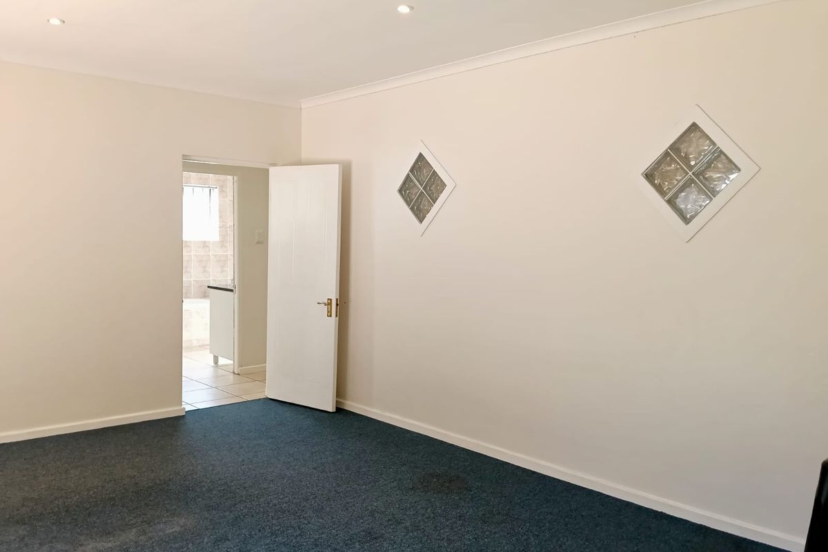 An empty bedroom featuring blue carpeting, recessed downlights, and decorative glass block wall inserts, with an adjoining bathroom visible through the open doorway.