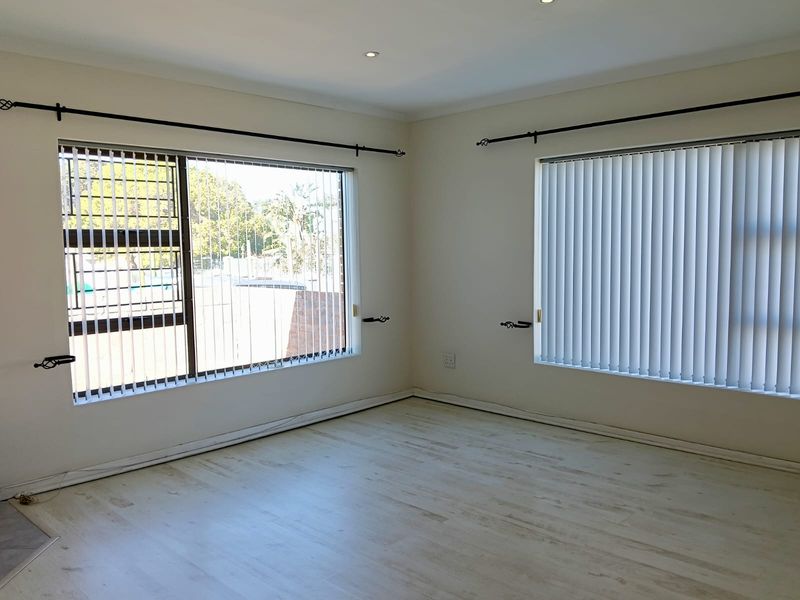 A bright, empty living room featuring light-toned laminate flooring, recessed downlights, and large windows fitted with vertical blinds and security bars.