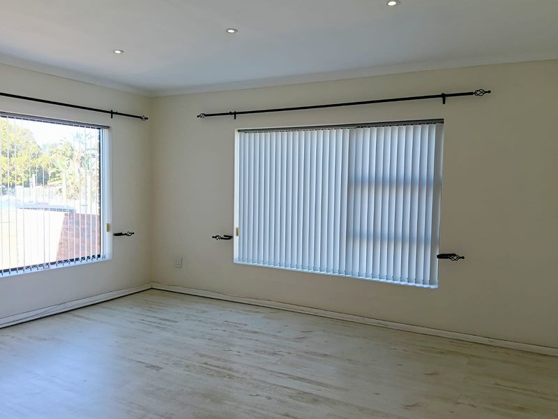 A bright, empty living room featuring light-toned laminate flooring, recessed downlighting, and vertical blinds on the windows.