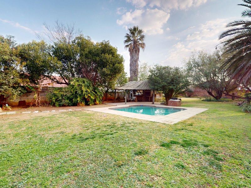 Spacious backyard featuring a swimming pool, paved surround, and a traditional thatched lapa, set within a mature garden with established trees.