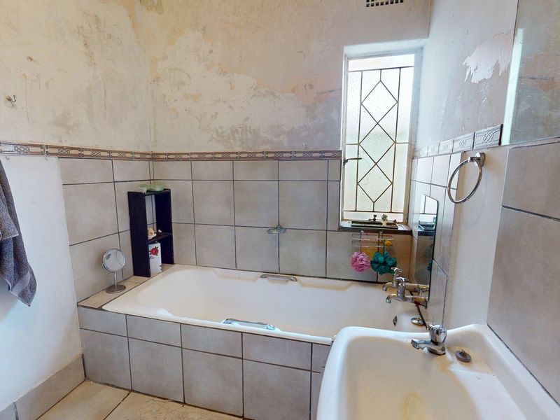 A dated bathroom featuring a built-in bathtub, pedestal basin, and leaded glass window, currently requiring significant wall repairs and cosmetic updates.