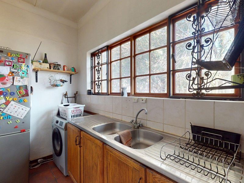 A functional scullery area featuring wooden cabinetry, a double stainless steel sink, tiled splashbacks, and timber-framed windows with security bars.