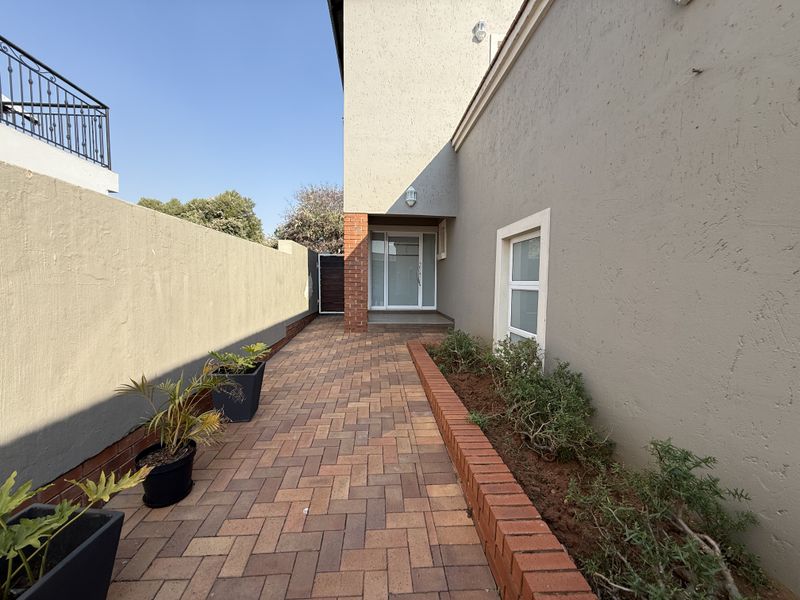 A well-maintained side walkway featuring brick paving, a raised brick planter bed, and a secure boundary wall leading to a glass entrance door.