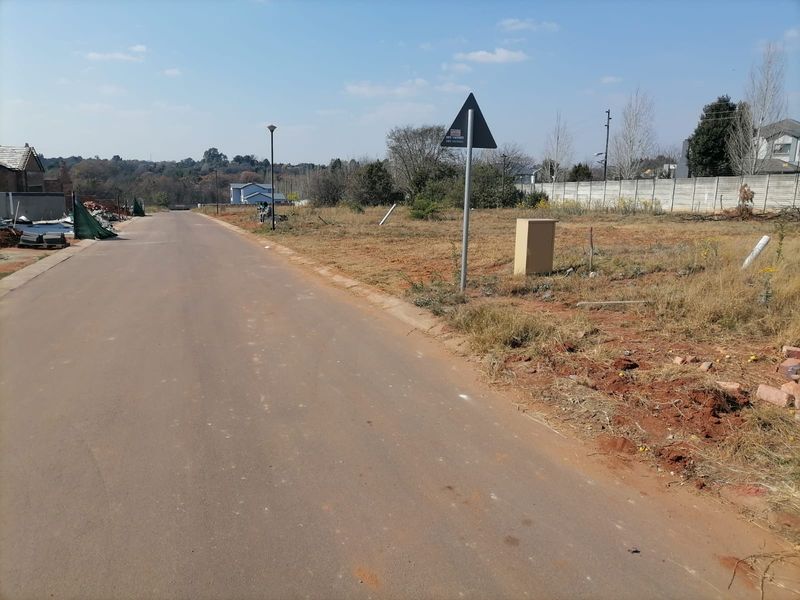 A street-level view of a vacant residential plot featuring an asphalt road, utility connection point, and surrounding boundary walling.