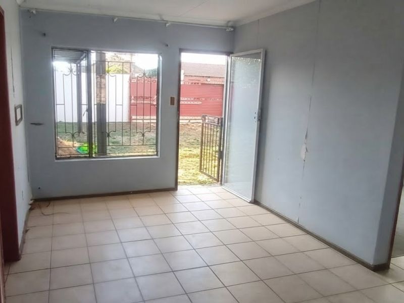 An empty living room featuring tiled flooring, security gate access to the exterior, and window security bars, requiring minor cosmetic wall repairs.