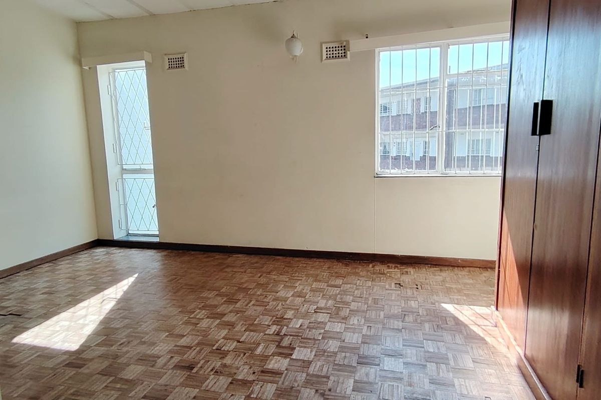 A spacious, empty bedroom featuring original parquet flooring, a built-in wooden wardrobe, and security bars on the windows.