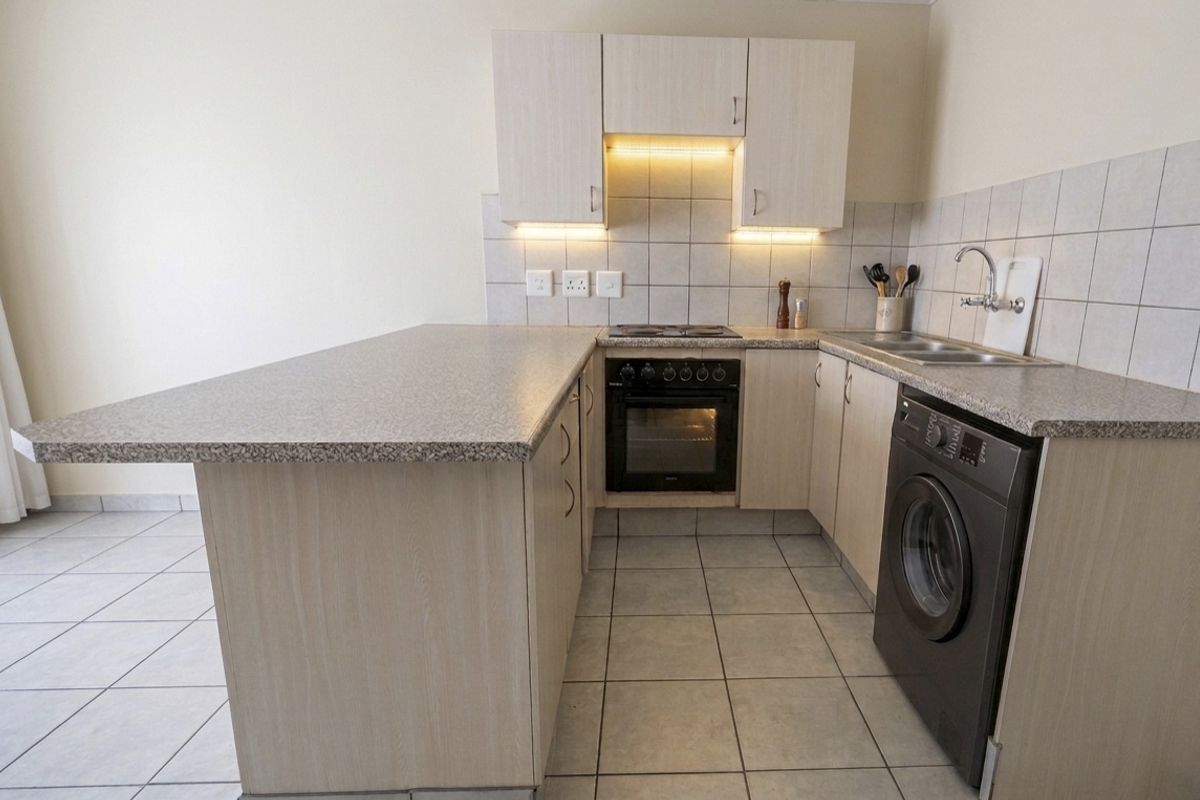 Compact kitchen featuring light wood-finish cabinetry, granite-look laminate countertops, built-in oven with hob, and tiled flooring with integrated laundry appliance space.