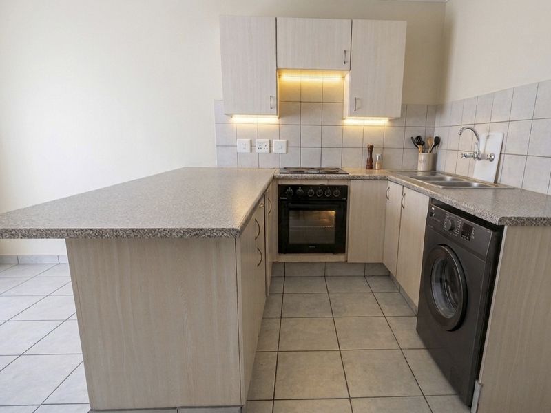 Compact kitchen featuring light wood-finish cabinetry, granite-look laminate countertops, built-in oven with hob, and tiled flooring with integrated laundry appliance space.