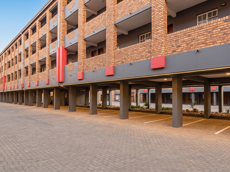 A modern apartment building exterior featuring a brick facade, covered ground-level parking, and paved driveway access.