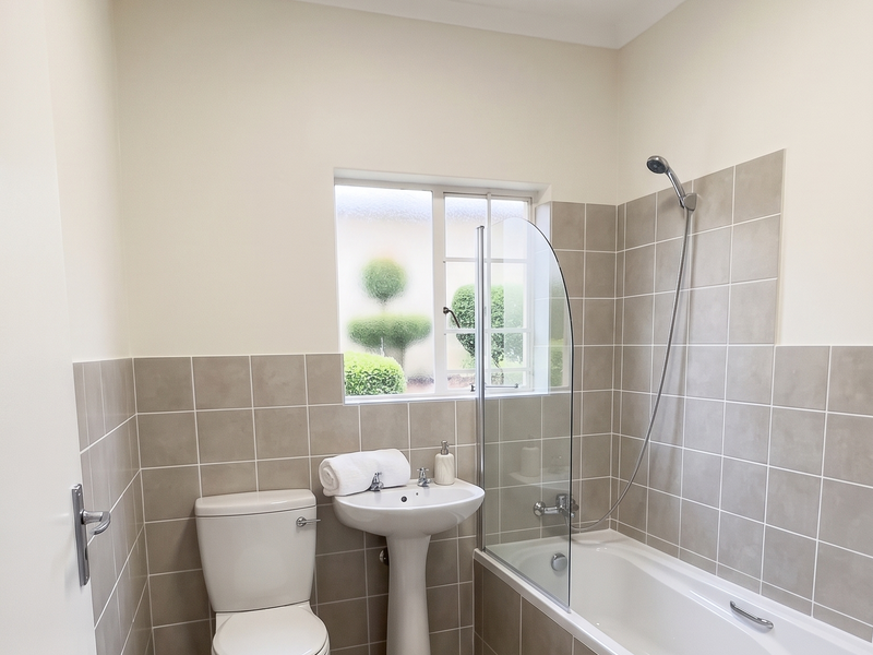 A clean, well-maintained bathroom featuring a pedestal basin, toilet, and a bathtub with a glass shower screen and neutral wall tiling.