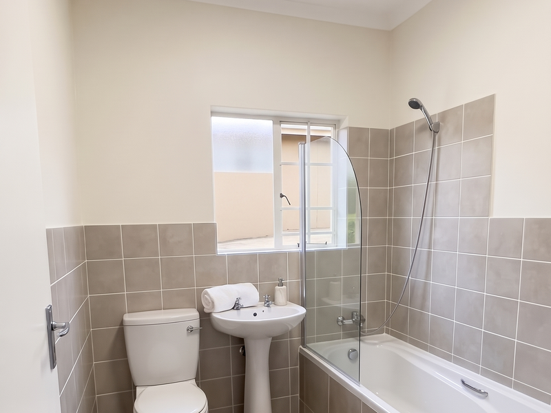A clean, modern bathroom featuring a pedestal basin, toilet, and a bathtub with a glass shower screen and neutral wall tiling.