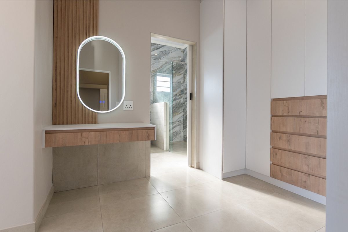 Modern bedroom dressing area featuring a sleek built-in vanity with LED mirror, extensive built-in cabinetry, and neutral tiled flooring, leading into an ensuite bathroom.