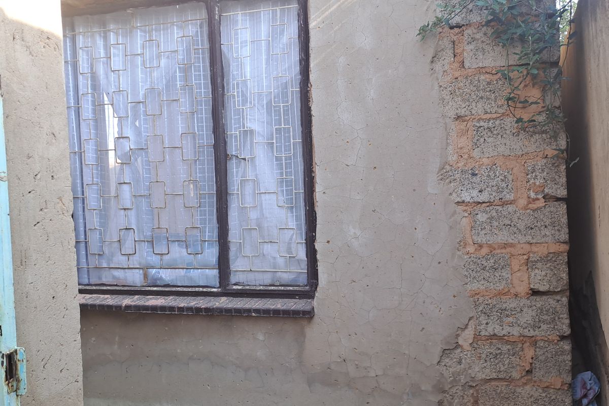 Exterior view of a side passage showing weathered plaster walls, exposed brickwork, and a window with security bars.