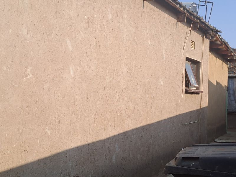 Side view of the property showing a plastered exterior wall, casement window, and corrugated metal roofing with a solar water heater installation.