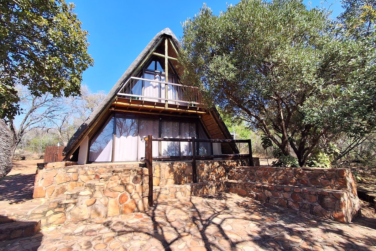 A charming A-frame house featuring a thatched roof, stone-clad exterior, and a private timber balcony overlooking a natural garden setting.