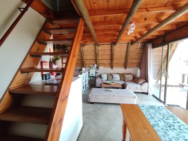 Open-plan living area featuring exposed timber beams, a thatched roof, concrete floors, and a timber staircase leading to an upper level.