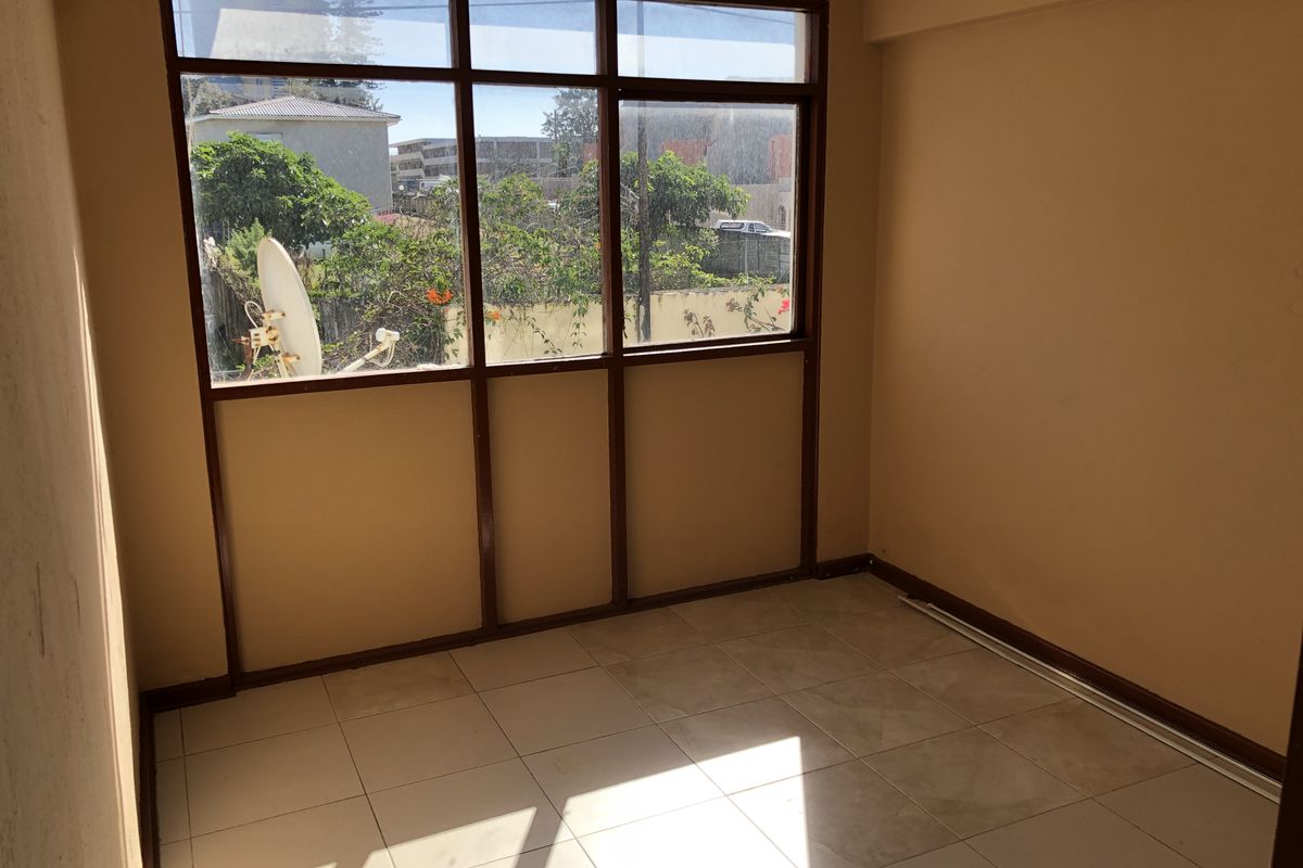 An empty bedroom featuring tiled flooring and large windows with aluminium frames, offering natural light and a view of the surrounding area.