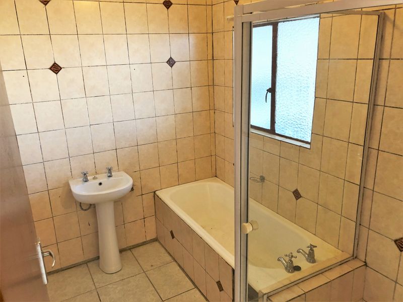 A dated bathroom featuring a pedestal basin, built-in bathtub with shower screen, and full-height wall tiling, offering potential for a modern renovation.