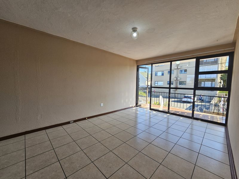 An empty, sunlit living area featuring neutral tiled flooring and large glass sliding doors that provide access to an outdoor balcony space.