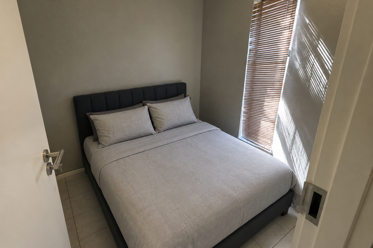 A compact bedroom featuring neutral wall tones, tiled flooring, and a window fitted with venetian blinds, providing a clean and functional living space.