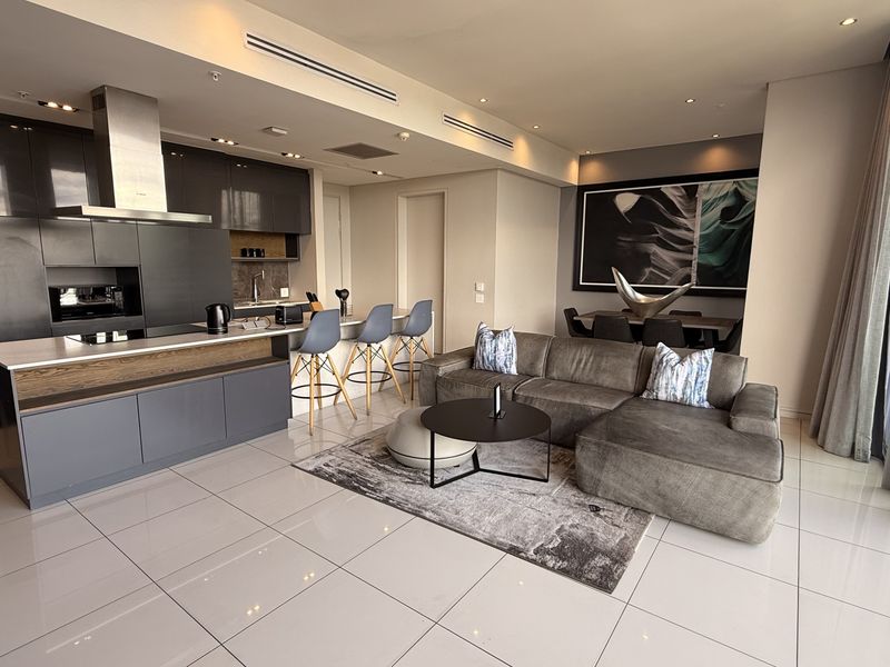 Modern open-plan penthouse living area featuring a sleek kitchen with integrated appliances, tiled floors, and floor-to-ceiling glass doors leading to a balcony.