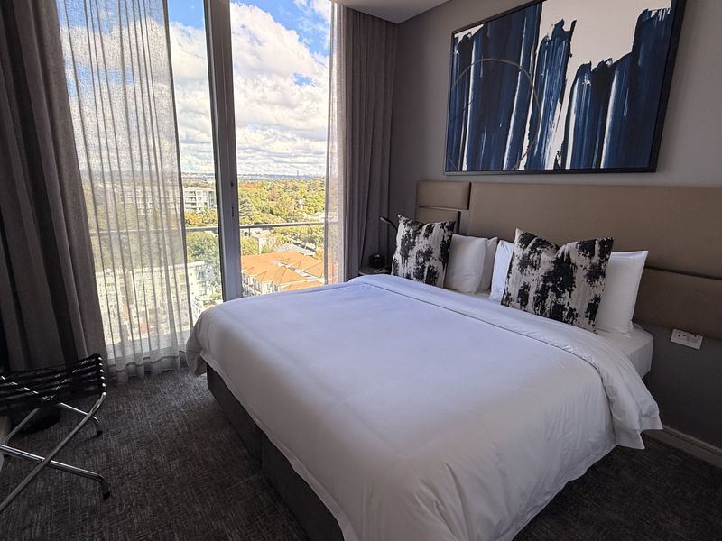 A modern penthouse bedroom featuring floor-to-ceiling windows with expansive city views, a custom upholstered headboard, and high-quality finishes.