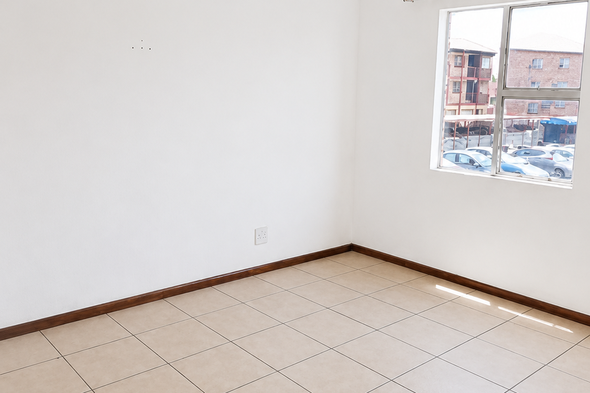 A bright, empty bedroom featuring neutral tiled flooring, white walls, and a large window providing natural light and views of the complex parking area.