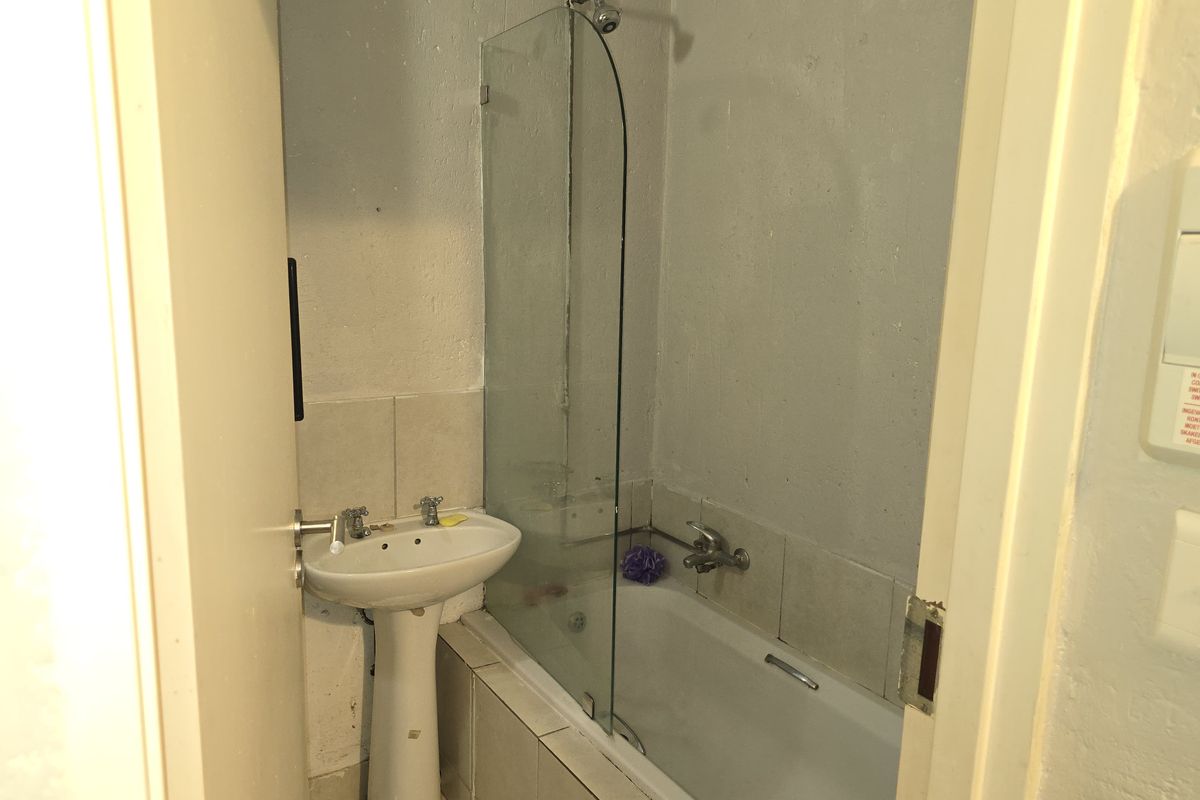 A compact bathroom featuring a pedestal basin, a bathtub with a glass shower screen, and tiled splashbacks, requiring cosmetic updates to improve its condition.