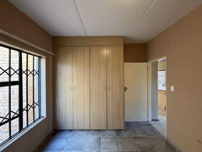 A functional bedroom featuring built-in wooden cupboards, tiled flooring, and security bars on the window, offering a clean and neutral space.