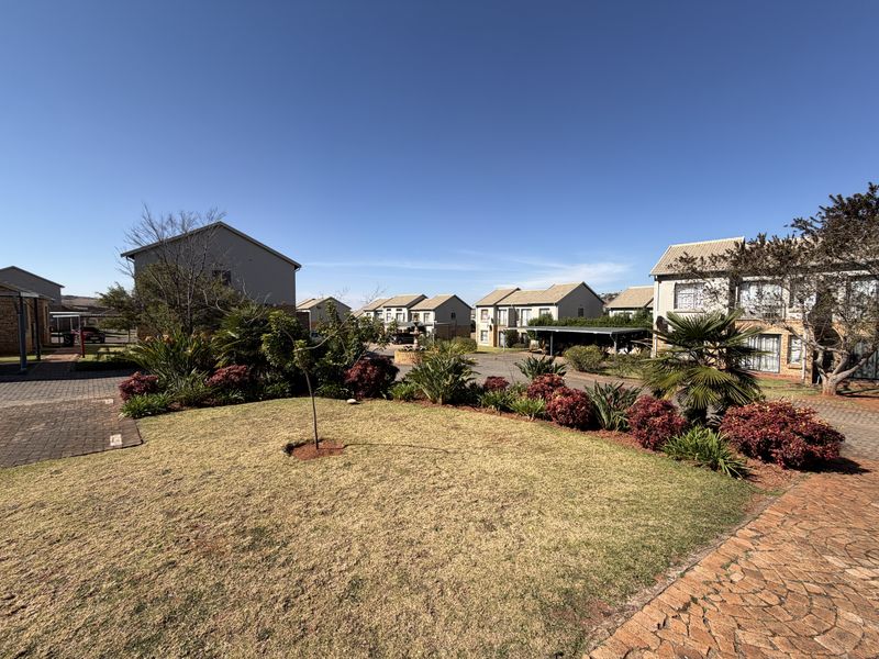 A view of the apartment complex grounds featuring a well-maintained lawn, landscaped garden beds, paved walkways, and covered carport parking areas.