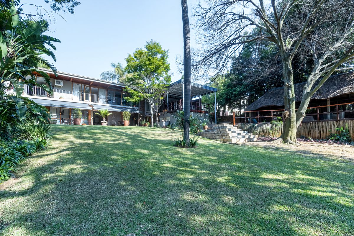 Spacious rear garden featuring a large lawn, mature trees, stone steps, and a multi-level dwelling with a balcony and separate thatched outdoor entertainment area.